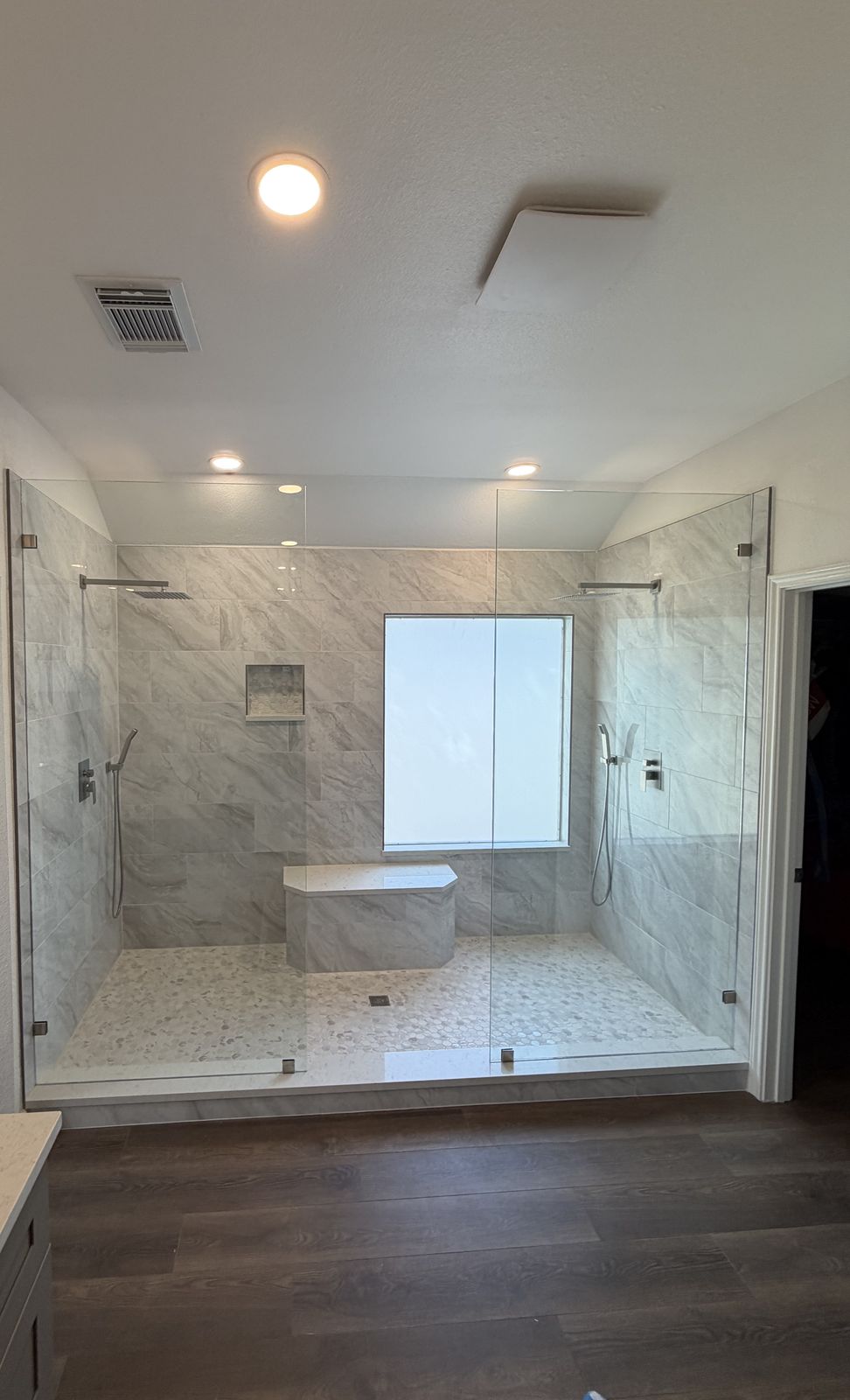 After: one large walk-in shower with dual showerheads, a bench and a niche, with two glass panels and no door