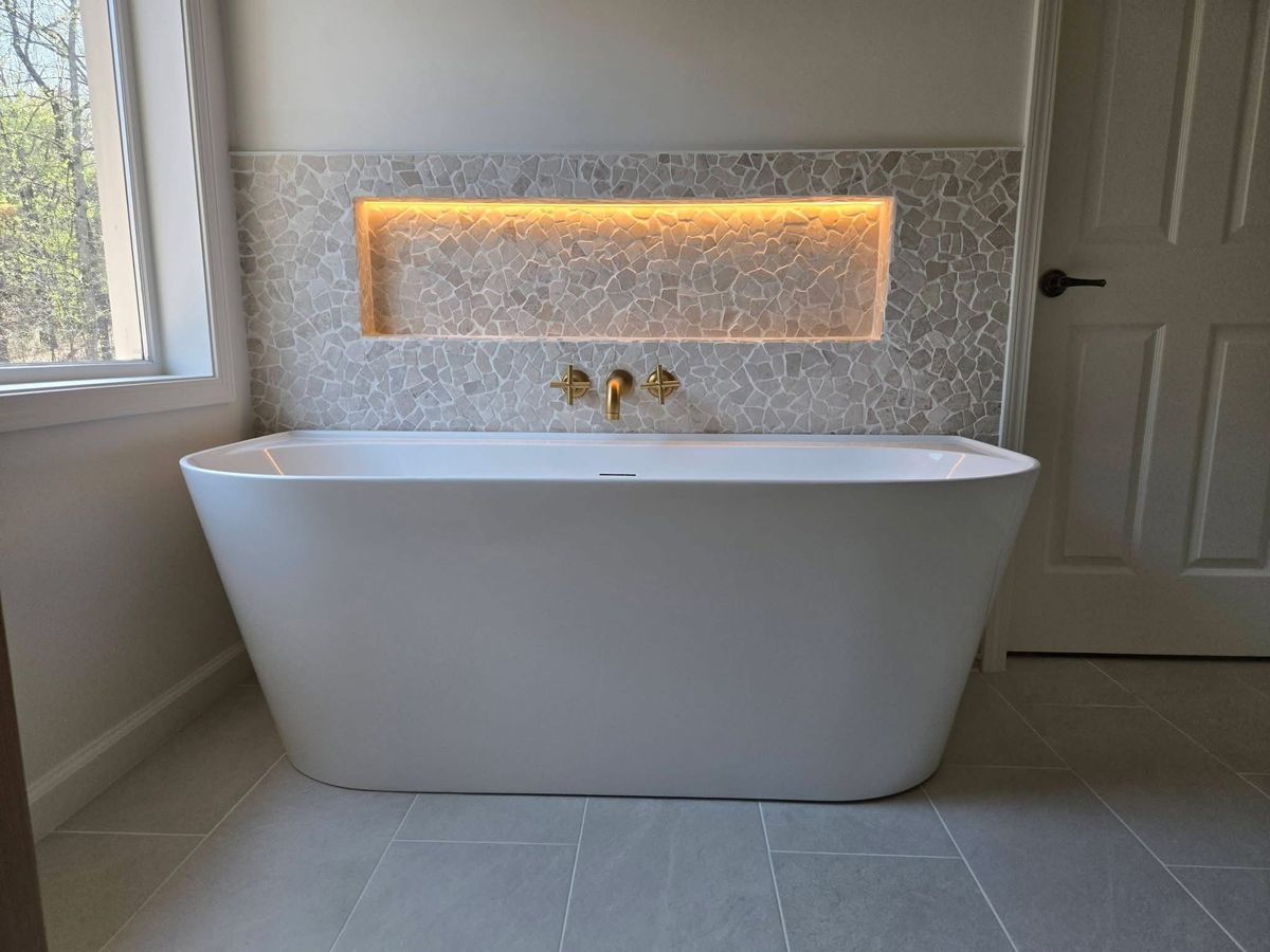 Freestanding soaking tub with brass wall-mounted faucet in front of a backlit stone mosaic niche, San Antonio bathroom remodel