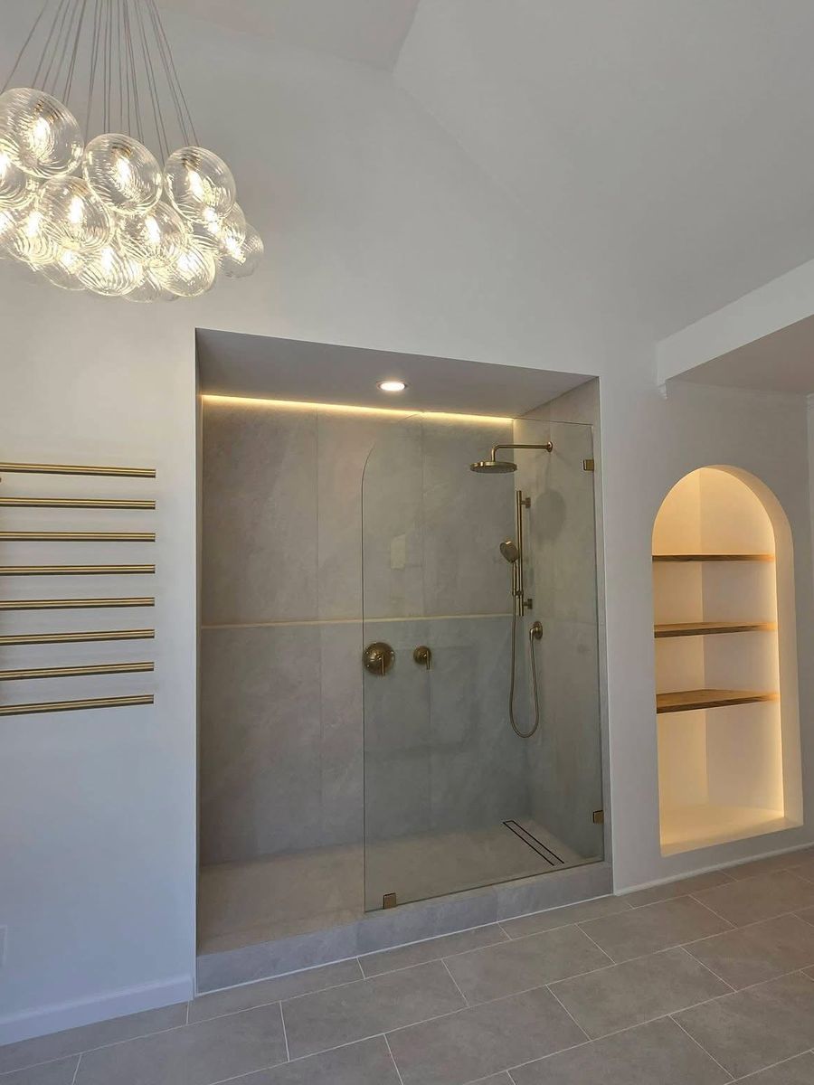Walk-in glass shower with brass fixtures and a linear drain, next to a lit arched shelving niche and a brass towel warmer