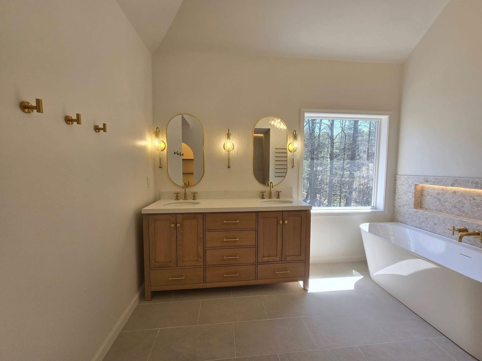 Primary bathroom remodel with a natural oak double vanity, oval brass mirrors and sconces, and a freestanding tub beside a lit stone niche