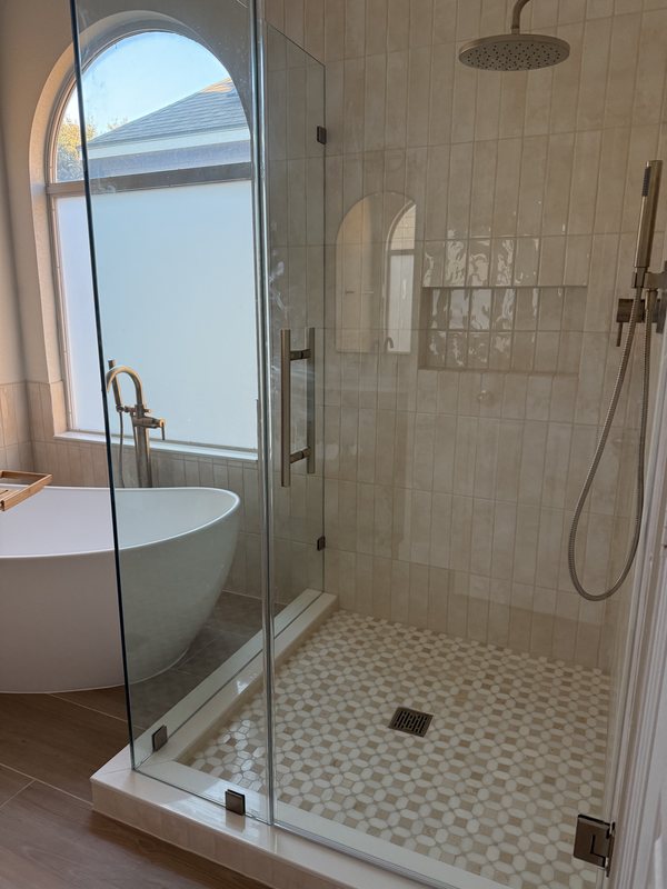 Frameless glass walk-in shower with cream tile, a mosaic floor and a built-in niche beside a freestanding tub under an arched window, San Antonio bathroom remodel
