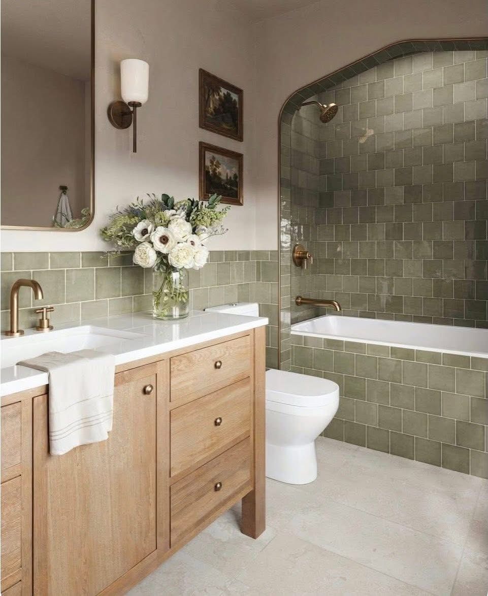 Bathroom with sage green tile, an arched tub-and-shower alcove, brushed brass fixtures and an oak vanity with a white top