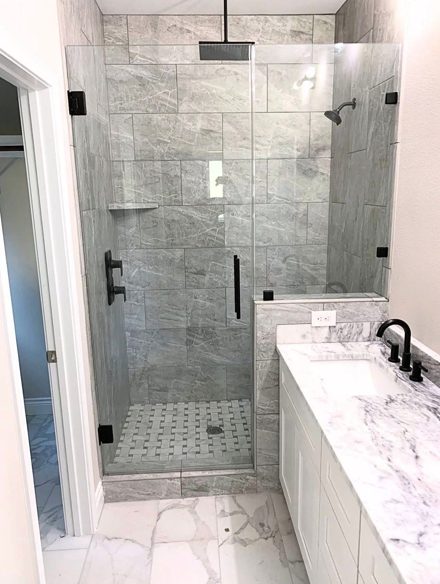 Frameless glass walk-in shower with large gray marble-look tile, basketweave floor and matte black fixtures beside a marble vanity