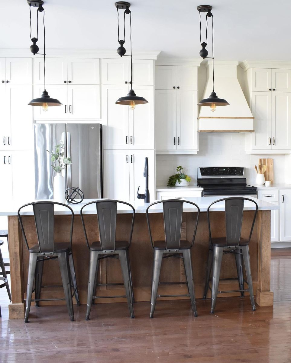 White shaker kitchen with a range hood trimmed in wood, stainless appliances and pulley pendants over a wood island